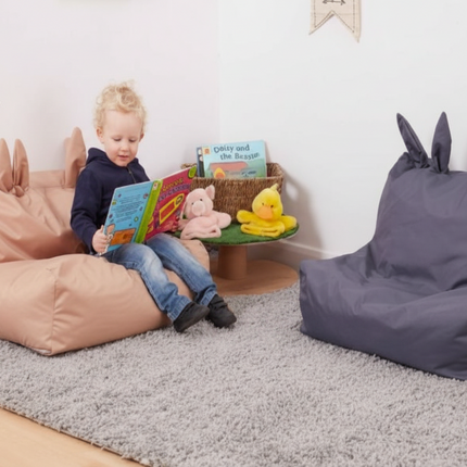 Child reading a book on a pink bean bag chair with a gray rug and blue bean bag chair in the background.