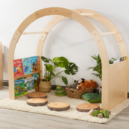 Children's play area with wooden arch, books, toys, and plants on a light wood floor.