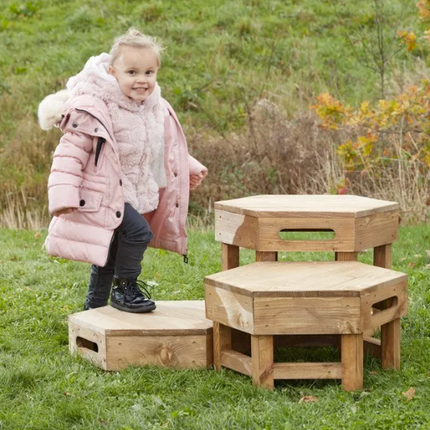 Child in a pink coat standing on wooden hexagonal play structures in a grassy outdoor area.