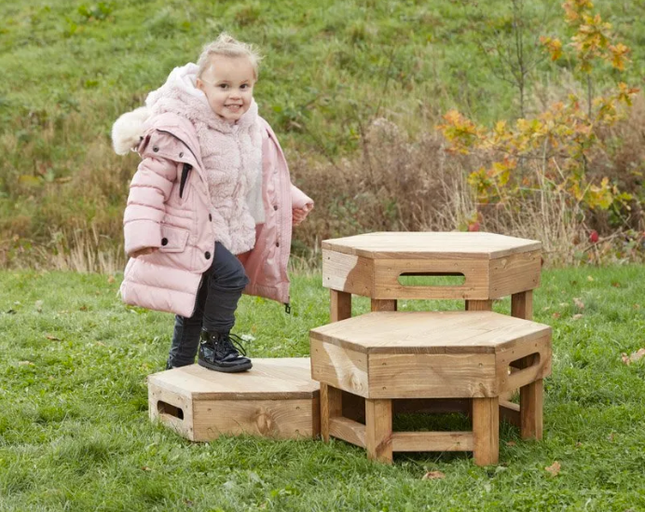 Child in a pink coat standing on wooden hexagonal play structures in a grassy outdoor area.